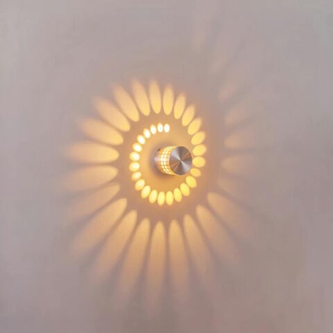 Applique LED circolare in alluminio argentato accesa, proietta una spirale di luce calda su una parete bianca