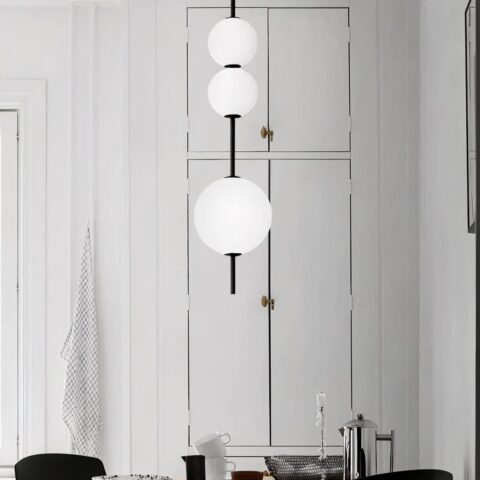 Lampadario suspendu à la verticale composé de trois boules blanches allumées glissées sur un axe. La boule de bas est plus grande que les deux petites en haut. Dans le fond, on voit un mur blanc et des portes de placard blanc.