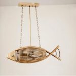 Lampadario en bois en forme de poisson, suspendu par deux chaines à chaque extrémité. Relier à un socle rectangulaire en bois. Derrière on voit un mur beige.