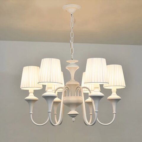 Lampadario blanc style baroque, en forme de chandelier avec 6 abat-jours blanc, les ampoules sont allumées. Suspendu par une chaine à un plafond blanc, on voit un bout de mur blanc dans le fond.