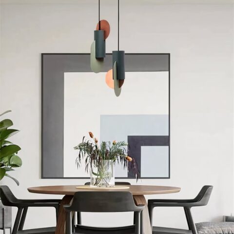Lampadarios design art-déco moderne, suspendus au-dessus d'une table ronde avec un vase avec des fleurs. Il y à trois chaises autour de la table, dans le fond un mur blanc et un grand tableau avec du gris, blanc, bleu et noir en forme de L.