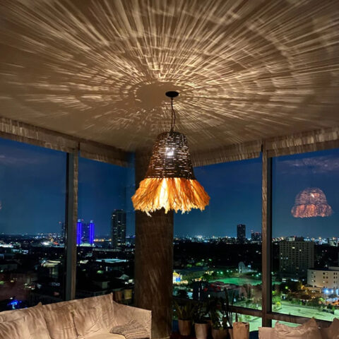 Lampadario paille et raphia tissé main, accroché au plafond et allumé, dans une pièce de vie type salon. Illumine le plafond de ses jeux d'ombres grâce au tissage du raphia et de la paille.