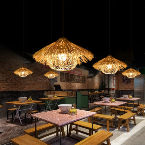 Lampadario paille naturelle style paillote exotique accrochée au mur et allumée, au dessus de tables de ce qui semble etre un restaurant et dans une ambiance de lumière tamisée.