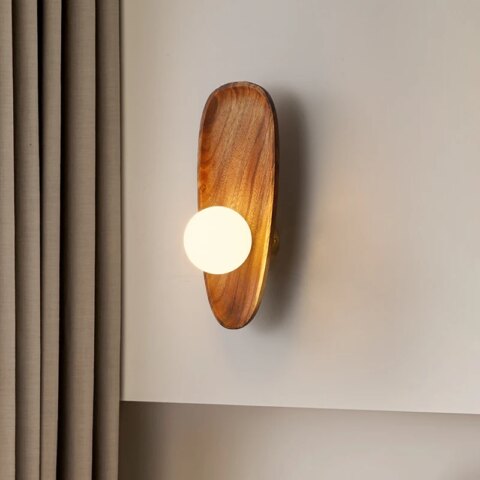 Applique murale en bois style coquillage avec sa perle lumineuse grand modèle, accrochée sur un mur blanc, à côté d'un rideau couleur taupe.