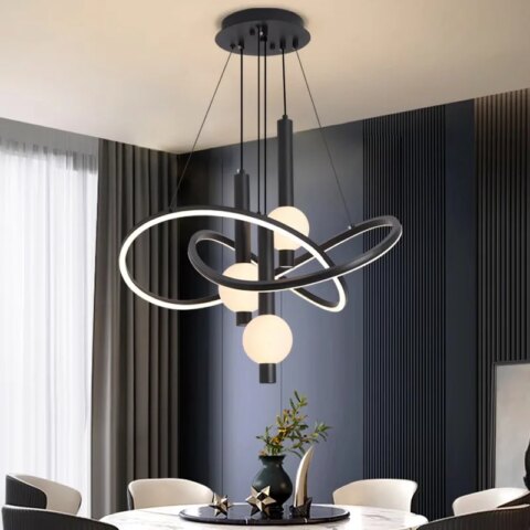 Lampadario noir avec support noir fixé au plafond d'où descendent des câbles tenant des structures arrondies entrelacées lumineuses et noires à l'extérieur. Au centre de ces structures, sont pendus des tubes noirs avec une boule lumineuse allumée. Fixé au-dessus d'une table blanche ronde style marbre, avec plateau noir rond, un vase dessus et des objets de décoration. 6 fauteuils autour de la table,à gauche une baie vitrée avec un rideau gris et le mur en face est bleu canard avec des rainures verticales.