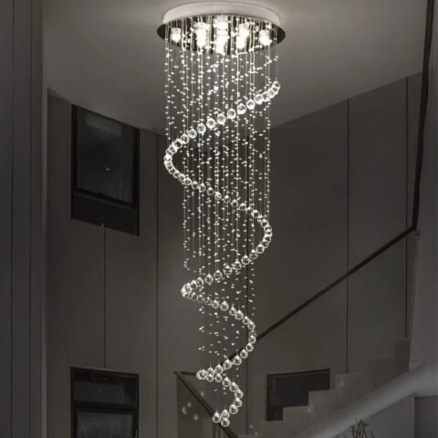Lampadario plafonnier en forme de spirale de suspensions en gouttes d'eau. Lumière blanche allumée par plusieurs LED dans le support rond argenté fixé au plafond. Derrière, on voit un escalier avec rambarde noire et des tableaux sur le mur.