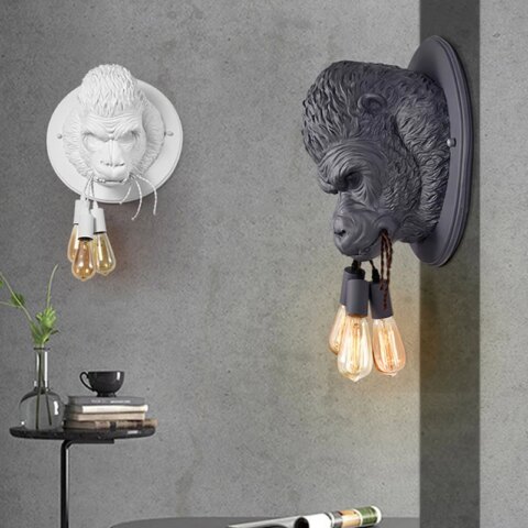 Deux appliques murales rondes avec la tête d'un gorille en 3D tenant dans sa bouche 3 cordes avec une ampoule suspendue à chacune. Sur le mur gris en face, l'applique est blanche, sur le mur gris de droite, elle est noire. Sous la blanche, une table ronde noire avec un seul pied fin. Dessus un vase transparent avec un feuillage, des livres et une tasse sur la pile de livres.
