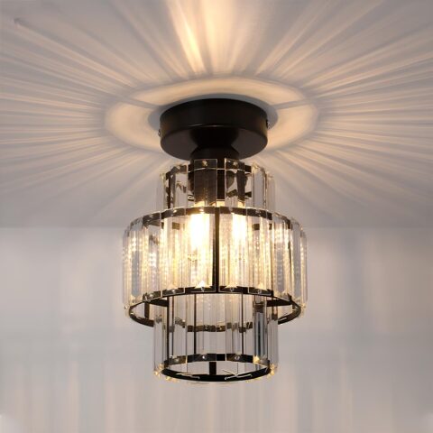 Lampadario plafonnier de forme cylindrique avec 3 rangées de lamelles style cristal. Le support u plafond est de couleur noire et les lamelles sont fixées sur des cercles noirs. Les ampoules sont allumées et diffusent des rayons au plafond autour du lustre.