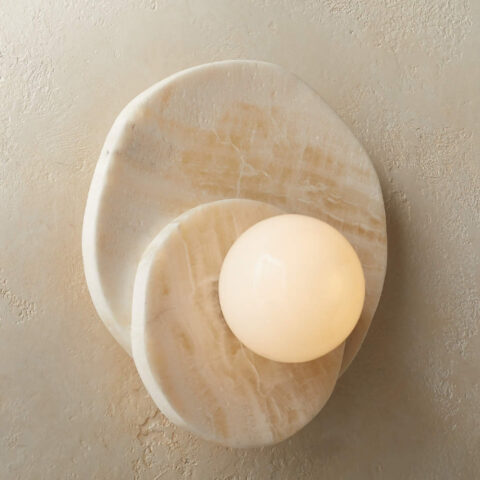 Applique murale circolare in pietra naturale di colore beige con due cerchi in pietra uno sull'altro. Al centro, una sfera luminosa. L'applique è fissata su una parete beige.